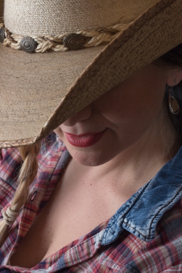 Close up photographic portrait of blonde model, hair in plaits, wearing cowboy hat, face partially obscured by brim of hat.